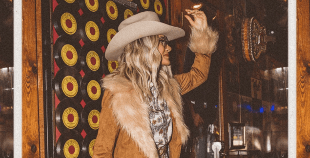 Female creator posing for a picture in a tan cowgirl hat and light brown coat in a bar.