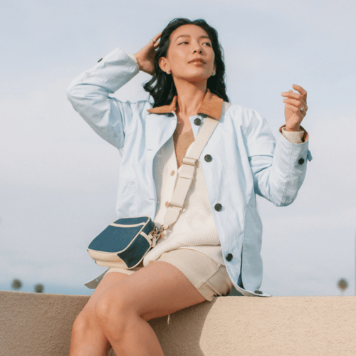 Woman with dark hair in a blue jacket, cream shorts, and crossbody bag posing on a ledge