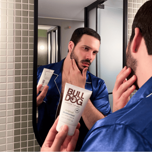 Man wearing blue silk pajamas promoting skincare product as he looks into the mirror applying moisturizer