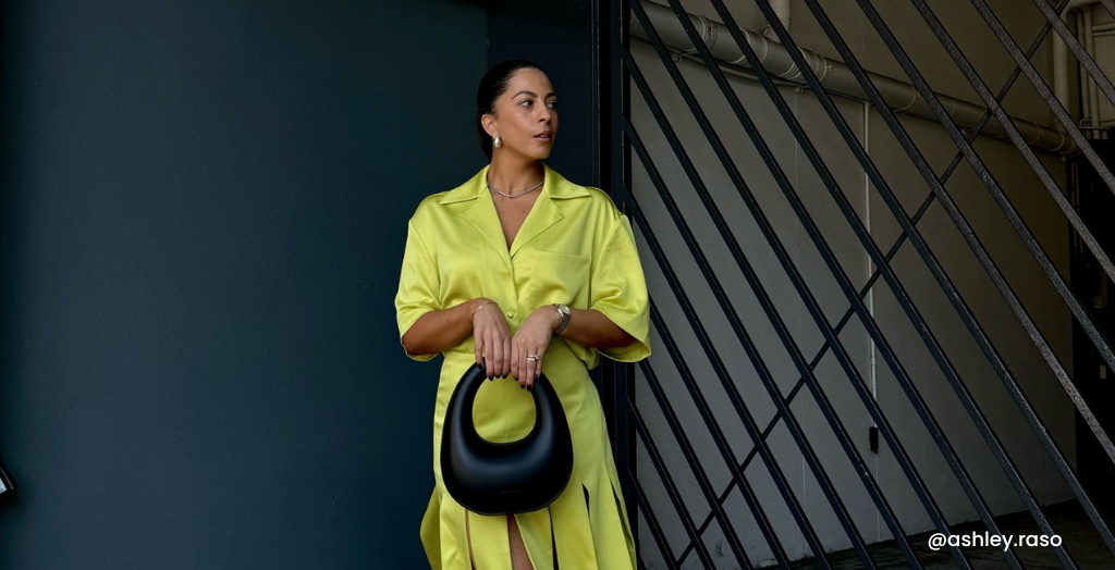 Creator @ashley.raso poses for a picture while wearing a silk dress in chartreuse and holding a black leather purse.