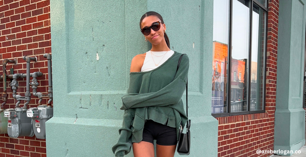 Creator @amberlogan.co in a green off-shoulder sweater, white tank, and black shorts posing casually on the street.