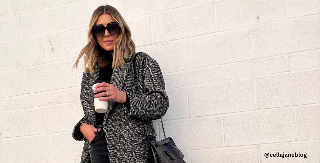 Content creator cellajaneblog posing for a photo wearing in dark shades and a black trench coat while holding a cup of coffee.