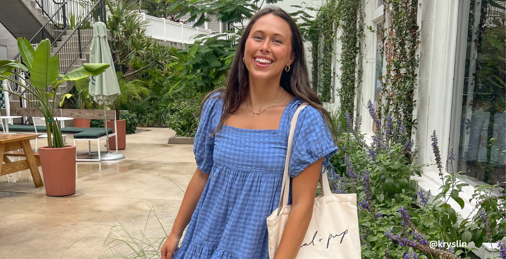 Creator @kryslin on Instagram smiling for a photo in a blue dress holding a cream over-the-shoulder bag